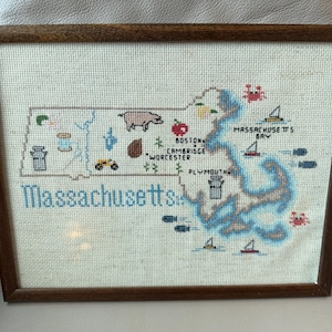 May include: Cross-stitch map of Massachusetts in a wooden frame. The map features embroidered icons representing various locations and landmarks, including a pig, apple, and sailboats. The word "Massachusetts" is stitched in blue.