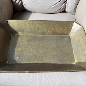 May include: A rectangular brass tray with angled, sloping sides and a flat base. The metal surface features a weathered, aged patina with visible variations in tone and texture, showing a metallic gold and brownish finish throughout the piece.