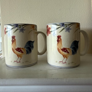 May include: Two cream-colored ceramic mugs featuring a repeating illustration of a rooster with a red comb, golden-brown neck feathers, and dark blue tail feathers. The design includes delicate floral accents in blue and orange at the top.