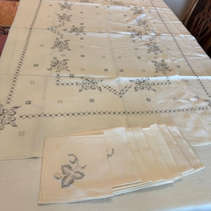 May include: Cream-colored tablecloth and matching napkins with a floral embroidery design in gray. The tablecloth is rectangular and covers a table. A stack of napkins is visible on the table.