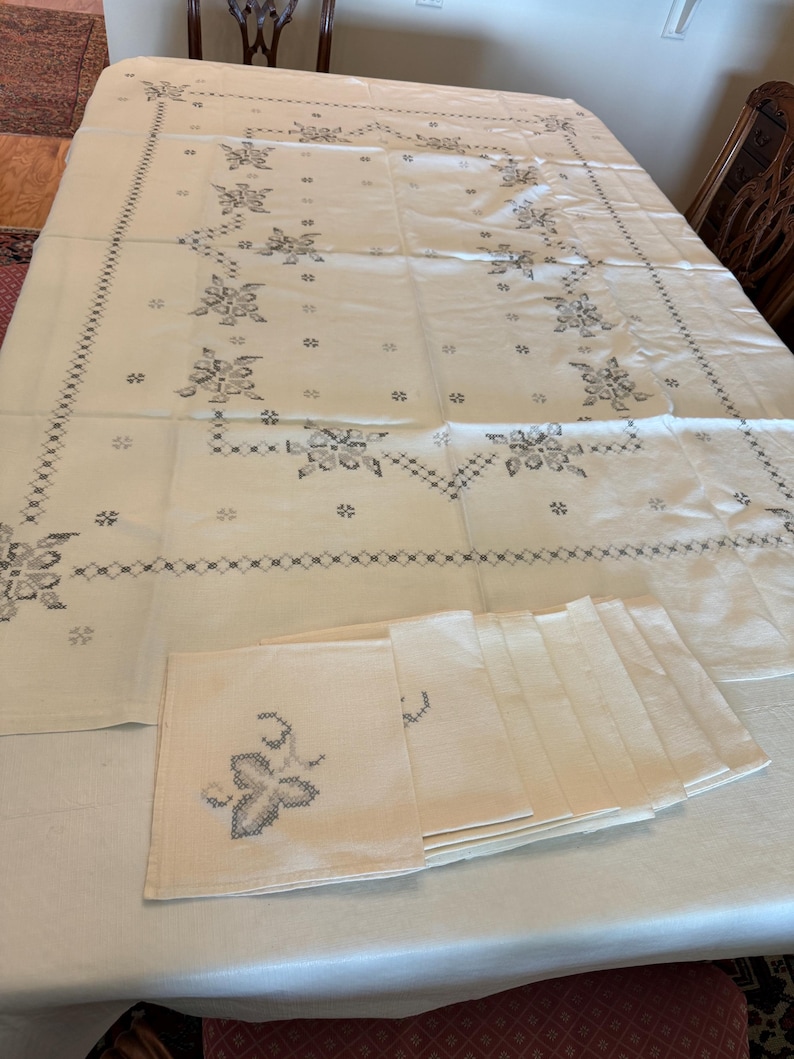May include: Cream-colored tablecloth and matching napkins with a floral embroidery design in gray. The tablecloth is rectangular and covers a table. A stack of napkins is visible on the table.