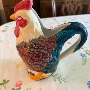 May include: A ceramic rooster pitcher with a red comb, yellow beak, and detailed feather patterns in shades of red, blue, and green. The handle is blue and white. The pitcher is on a floral tablecloth.