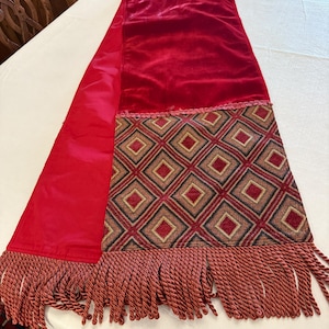 May include: A red velvet table runner with a patterned diamond design and fringe. The runner features a rich red color with a textured velvet top and a decorative diamond pattern in shades of red, brown, and gold.