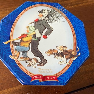 Könnte beinhalten: Achteckige blaue Dose mit einer Illustration von Norman Rockwell, die eine Person mit einem Weihnachtsbaum, ein Kind mit einer Axt und Hunde zeigt. Die Illustration befindet sich in einem weißen Oval, mit dem Text "Norman Rockwell CIRCA 1949".