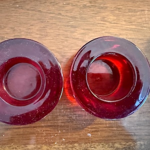Könnte beinhalten: Zwei rubinrote Kerzenhalter aus Glas. Jeder Kerzenhalter hat einen kreisförmigen Sockel mit einer zentralen Vertiefung zur Aufnahme einer Kerze. Das Glas ist transparent, so dass Licht hindurchscheinen kann. Die Kerzenhalter stehen auf einer Holzoberfläche.