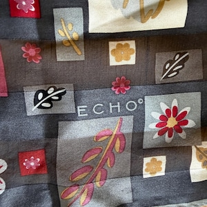Könnte beinhalten: Ein dunkelgrauer Schal mit einem sich wiederholenden Muster aus rechteckigen Blöcken, die verschiedene botanische Illustrationen enthalten, darunter rote, gelbe und weiße Blumen und Blätter. Das Wort ECHO ist in weißen Großbuchstaben in der Mitte aufgedruckt.
