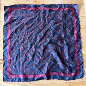 May include: A square silk scarf featuring a dark blue and magenta paisley pattern throughout the center. The scarf is framed by a solid magenta border and a thin outer edge decorated with a repeating blue and gold geometric motif.