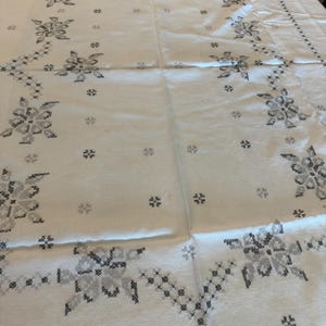 May include: A white tablecloth with a floral cross-stitch pattern in shades of gray. The design features large floral motifs and smaller, scattered cross-stitch elements, creating a delicate and detailed aesthetic. The edges are also decorated with a cross-stitch border.
