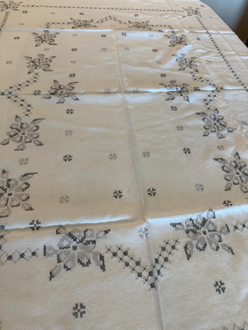 May include: A white tablecloth with a floral cross-stitch pattern in shades of gray. The design features large floral motifs and smaller, scattered cross-stitch elements, creating a delicate and detailed aesthetic. The edges are also decorated with a cross-stitch border.