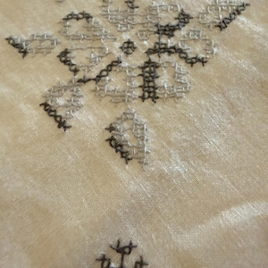 May include: Close-up of a cream-colored fabric with intricate cross-stitch embroidery. The embroidery features a floral design in shades of gray and black, creating a textured and detailed pattern. The fabric has a slightly wrinkled appearance.