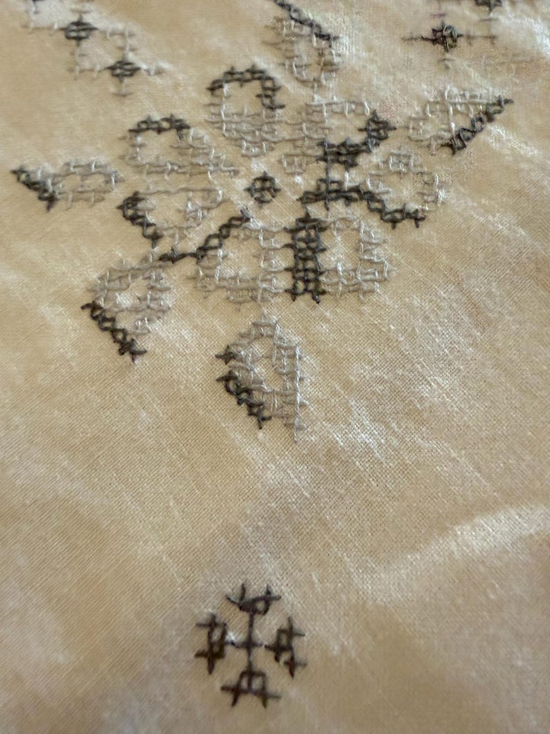 May include: Close-up of a cream-colored fabric with intricate cross-stitch embroidery. The embroidery features a floral design in shades of gray and black, creating a textured and detailed pattern. The fabric has a slightly wrinkled appearance.