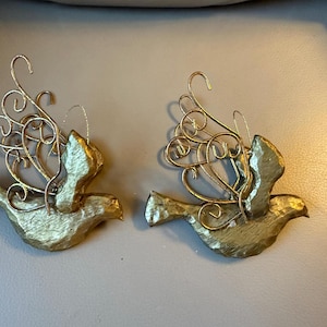 Pode incluir: Dois ornamentos de pomba dourados. Cada pomba tem asas e corpo texturizados, com delicados detalhes de arame em espiral para as asas. Os ornamentos são projetados para pendurar, sugerindo uso festivo ou decorativo.