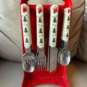 May include: A red plastic caddy holds a set of stainless steel flatware. The handles are white with a repeating pattern of green Christmas trees and red accents. The set includes a fork, knife, and two spoons.
