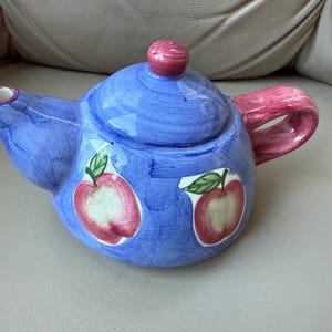 Könnte beinhalten: Eine blaue Keramik-Teekanne mit Deckel und Henkel. Die Teekanne ist mit roten Apfelmotiven und einem rosa Henkel verziert. Der Deckel hat einen rosa Knauf. Die Teekanne steht auf einer hellen Oberfläche.
