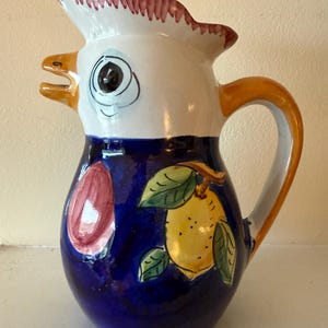 May include: A ceramic pitcher with a rooster head design. The pitcher features a white head with a red and white ruffled crest, a blue body with a painted lemon and fruit design, and an orange handle. The pitcher is a decorative kitchenware item.