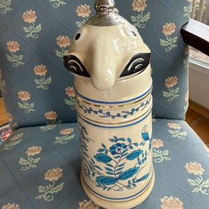 Peut inclure: Une chope à bière en céramique crème avec un visage fantaisiste sur le couvercle et un dessus en métal argenté. Le corps présente des motifs floraux et d'oiseaux bleus avec des bordures dorées. La chope mesure environ 25 cm de haut.