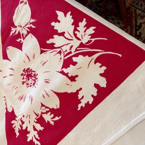 May include: A cream-coloured textile featuring a large, bold triangular section in deep red. The fabric is decorated with a white floral pattern, including a prominent central flower, detailed leaves, and a closed bud, all set against the red background.