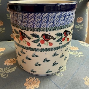 Può includere: Un portautensili cilindrico in ceramica con bordo blu scuro e corpo bianco. Il corpo presenta un motivo ripetuto di uccelli stilizzati, bacche e foglie in rosso, verde e nero su sfondo bianco.