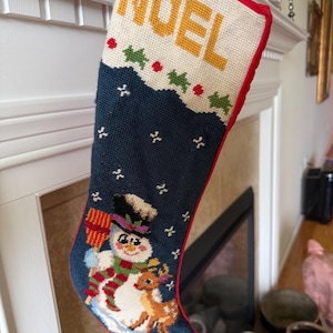May include: A Christmas stocking with the word "NOEL" in yellow letters. The stocking features a snowman and reindeer design on a navy blue background, with a red trim. The stocking is hanging on a white mantel.