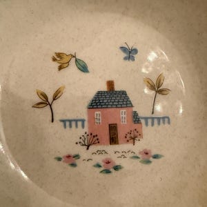 May include: A speckled off-white ceramic bowl featuring a central illustration of a pink house with a blue roof, a brown door, and white windows. The scene includes a yellow bird, a blue butterfly, green trees, a blue fence, and pink flowers.
