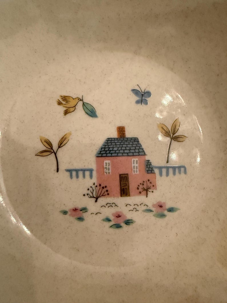 May include: A speckled off-white ceramic bowl featuring a central illustration of a pink house with a blue roof, a brown door, and white windows. The scene includes a yellow bird, a blue butterfly, green trees, a blue fence, and pink flowers.