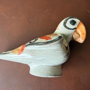 May include: A ceramic parrot figurine with a light gray body, black and orange accents, and a large orange beak. The bird has a round black eye and is perched on a small base. The figurine is a decorative item.