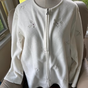 May include: A cream-colored fleece zip-up jacket featuring a round neckline and long sleeves. The fabric is decorated with an embroidered pattern of holly leaves and small red berries scattered across the front and sleeves. A silver zipper pull is present.