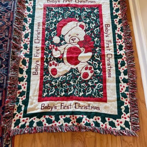 Puede incluir: Una manta tejida rectangular con un oso de peluche color crema en el centro, que lleva un gorro de Papá Noel rojo y sostiene un bastón de caramelo. El borde muestra un patrón de hojas de acebo verdes y bayas rojas con el texto Baby's First Christmas.