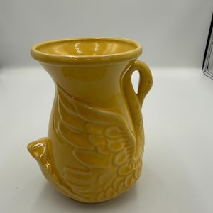 May include: A yellow ceramic swan vase. The vase features a handle and a detailed wing design. The swan's head forms the spout. The vase is a bright, solid yellow colour. A decorative item for the home.