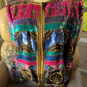 May include: A long-sleeve jacket featuring horizontal bands of sequins in vibrant shades of magenta, teal, blue, and black. The garment is accented with gold metallic embroidery along the front opening, neckline, and hem, with floral motifs.