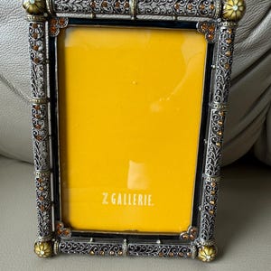 May include: A rectangular picture frame with an ornate silver-toned metal border featuring intricate filigree patterns and embedded amber-colored crystals. The corners are accented with rounded decorative caps, and the center displays a bright yellow insert with the white text Z GALLERIE printed at the bottom.