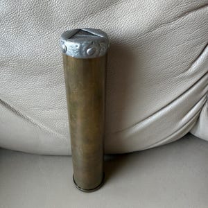 May include: A cylindrical brass tube with a weathered, dark gold patina. The top is capped with a textured, silver-coloured metal rim featuring a series of small, circular indentations around its circumference. The base is a simple, flat metal disc.