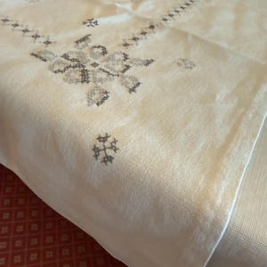 May include: Cream-colored linen tablecloth with intricate gray embroidered floral and geometric patterns. The embroidery is concentrated in the center and along the edges, creating a decorative border. The fabric is folded, showing its texture.