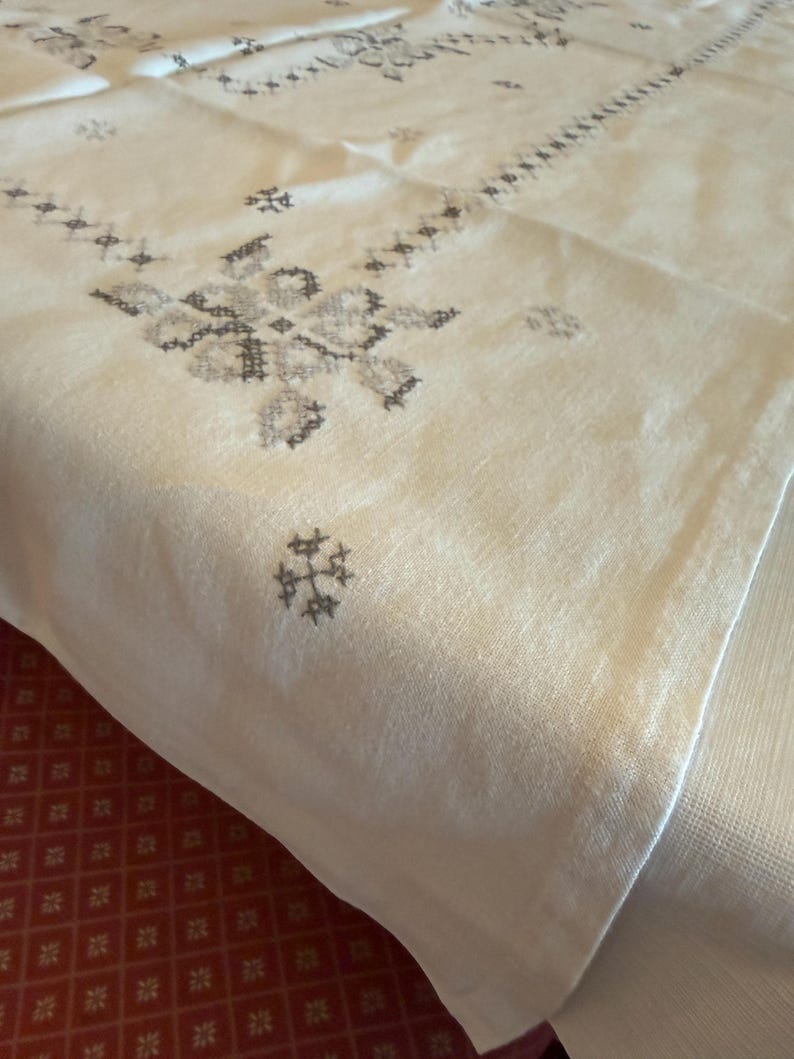 May include: Cream-colored linen tablecloth with intricate gray embroidered floral and geometric patterns. The embroidery is concentrated in the center and along the edges, creating a decorative border. The fabric is folded, showing its texture.