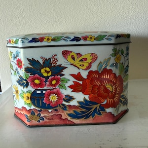 Puede incluir: Una caja de lata hexagonal vintage con fondo azul claro. La caja está decorada con coloridos diseños florales y de mariposas en rojo, amarillo y azul. La tapa y la base tienen un borde decorativo.
