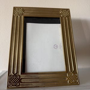 May include: A gold-colored picture frame with a rectangular shape. The frame has a decorative pattern at each corner and a black interior. The frame is resting on a white surface.