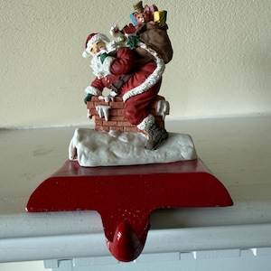 May include: A cast iron Christmas stocking holder featuring a painted Santa Claus figure climbing into a brick chimney. Santa wears a red suit with white trim and carries a brown sack filled with colorful gift boxes and a teddy bear.