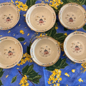May include: Six round ceramic bowls with a cream-colored interior and a central illustration of a small pink house with a blue roof, surrounded by green trees. The rim of each bowl features a delicate floral pattern with blue and yellow accents.