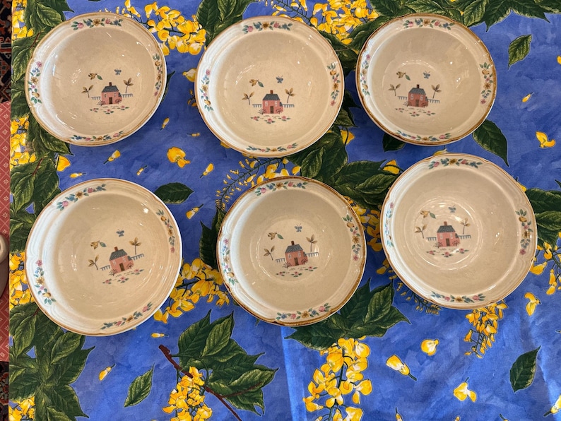 May include: Six round ceramic bowls with a cream-colored interior and a central illustration of a small pink house with a blue roof, surrounded by green trees. The rim of each bowl features a delicate floral pattern with blue and yellow accents.