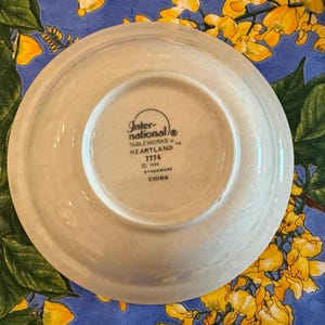 May include: A round, off-white stoneware saucer with a speckled texture. The underside features black printed text reading International Tableworks, Heartland, 7774, 1984, Stoneware, and China, centered within a circular indentation on the base.