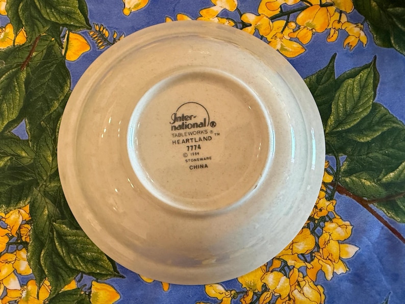May include: A round, off-white stoneware saucer with a speckled texture. The underside features black printed text reading International Tableworks, Heartland, 7774, 1984, Stoneware, and China, centered within a circular indentation on the base.