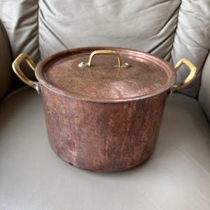 Puede incluir: Una olla de cobre con tapa y dos asas laterales, y un asa en la tapa. La olla tiene un aspecto deslustrado y envejecido, con un asa dorada. La olla está sobre una superficie gris claro.