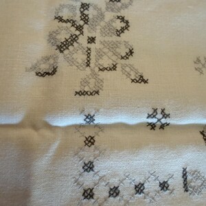 May include: Close-up of a cream-colored fabric with cross-stitch embroidery. The embroidery features floral and geometric patterns in shades of gray and black. The fabric has a slightly wrinkled texture.