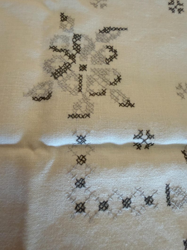 May include: Close-up of a cream-colored fabric with cross-stitch embroidery. The embroidery features floral and geometric patterns in shades of gray and black. The fabric has a slightly wrinkled texture.