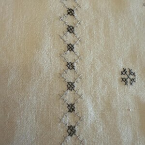May include: Close-up of off-white fabric with detailed embroidery. The embroidery features a repeating pattern of gray and black cross-stitch diamonds and floral motifs, creating a textured design. The fabric has a subtle, natural texture.