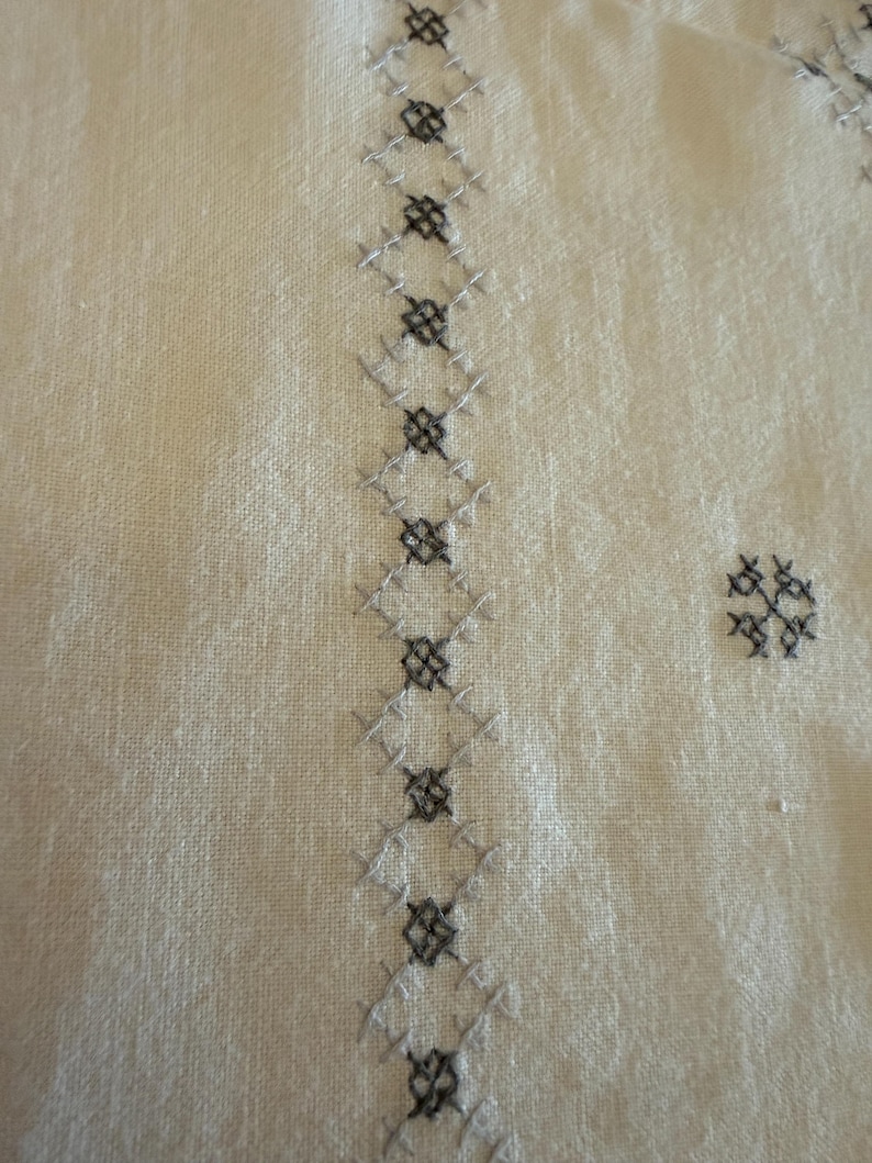 May include: Close-up of off-white fabric with detailed embroidery. The embroidery features a repeating pattern of gray and black cross-stitch diamonds and floral motifs, creating a textured design. The fabric has a subtle, natural texture.