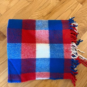 May include: A folded blanket with a bold plaid pattern in red, white, and blue. The blanket has a fringed edge with red and white tassels. The blanket is made of a soft, woven material and is folded neatly on a wooden surface.