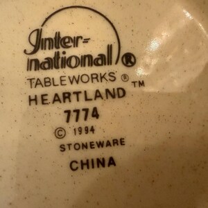 May include: A cream-colored stoneware base with dark brown speckles featuring the black printed text: International TABLEWORKS HEARTLAND 7774 &copy; 1994 STONEWARE CHINA. The text is arranged in a vertical stack with a curved line above the brand name.