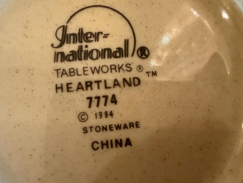 May include: A cream-colored stoneware base with dark brown speckles featuring the black printed text: International TABLEWORKS HEARTLAND 7774 &copy; 1994 STONEWARE CHINA. The text is arranged in a vertical stack with a curved line above the brand name.