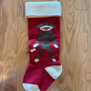 May include: A red knit Christmas stocking featuring a brown sock monkey graphic with white accents on the hands, feet, and mouth. The stocking has a wide cream-colored cuff at the top and a matching cream-colored heel and toe section.